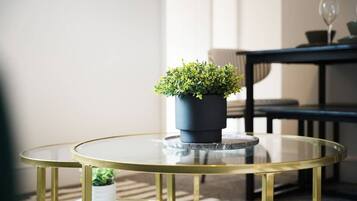 A round glass table with a black planter on top, a gold-framed chair, and a sofa in the background.