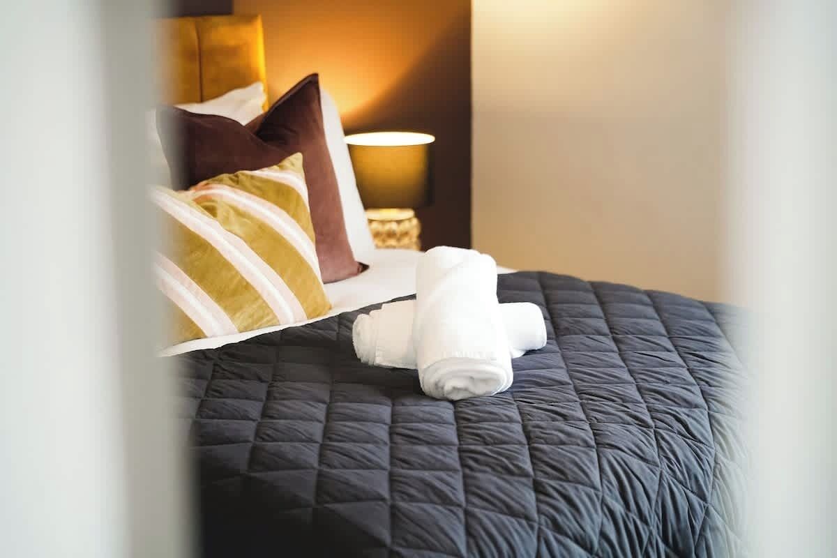 A neatly made bed with a dark quilt, a rolled white towel, and a brown pillow.