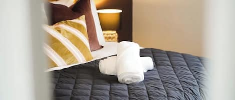 A neatly made bed with a dark quilt, a rolled white towel, and a brown pillow.