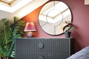 A room with a dark-colored dresser, a round mirror, a potted plant, and a lamp.