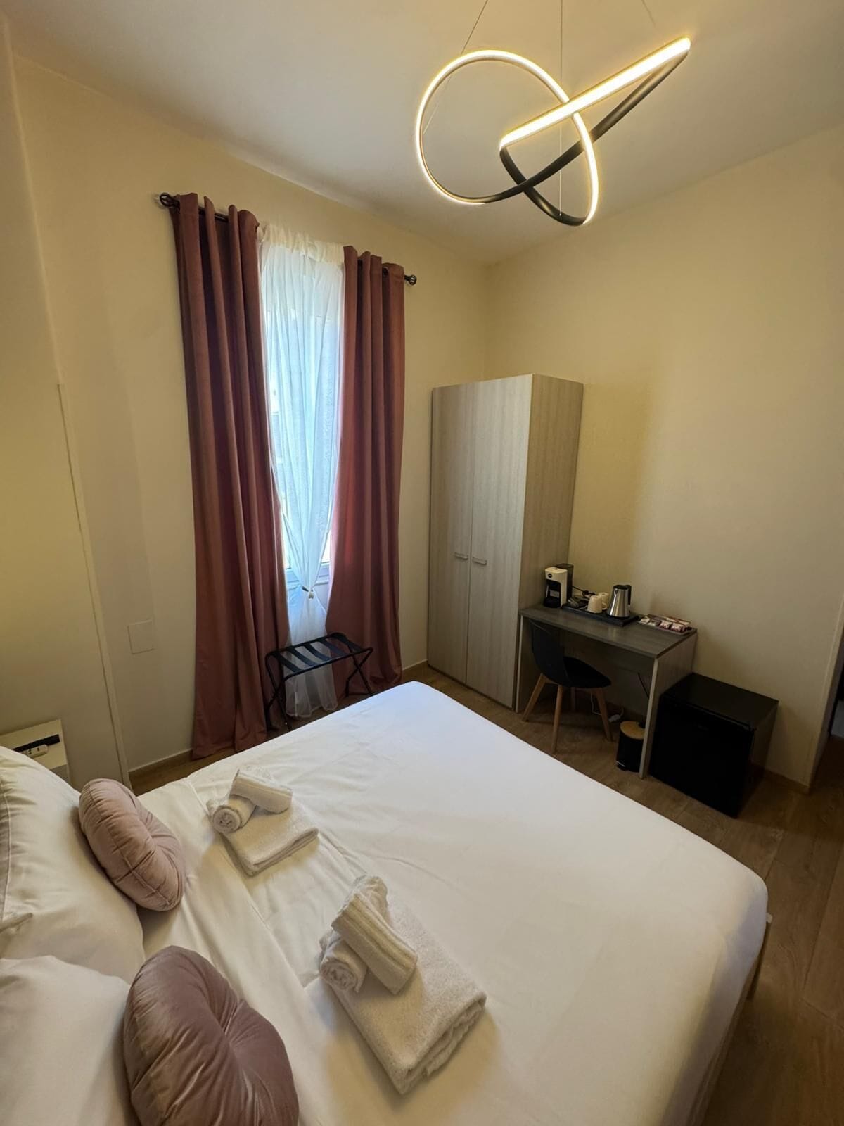 A hotel room with a bed, a desk, a wardrobe, and a modern light fixture.