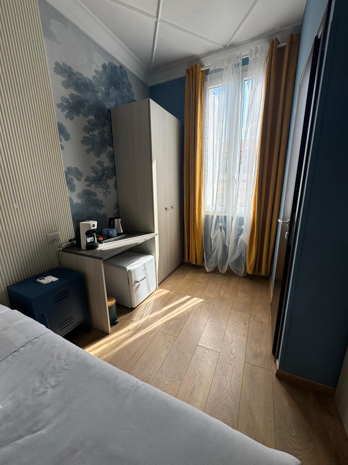 Habitación de hotel con cama, escritorio, armario, radiador y ventana con cortinas.