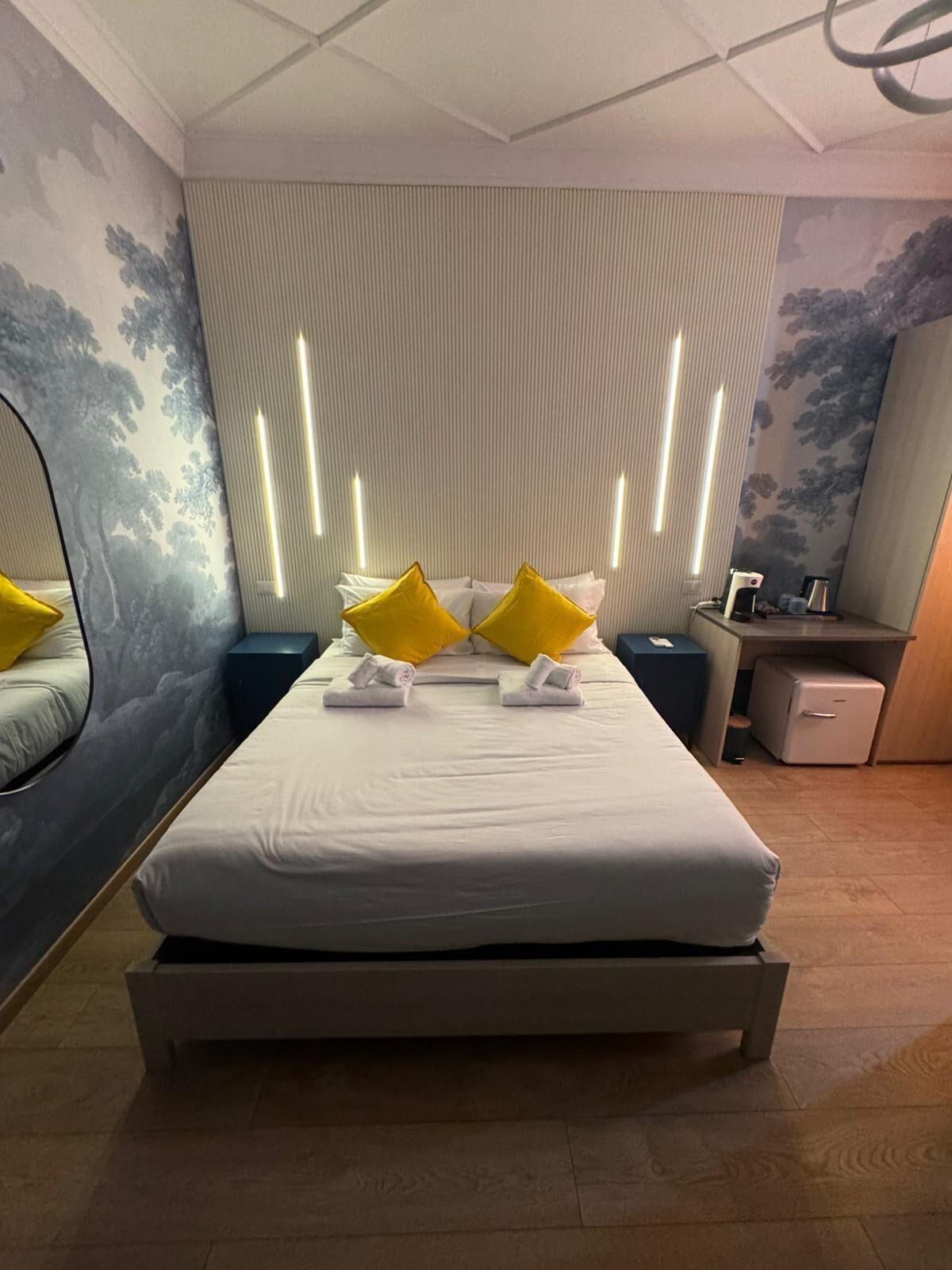 Une chambre d’hôtel moderne avec un grand lit, deux tables de chevet, un petit bureau et un réfrigérateur compact.