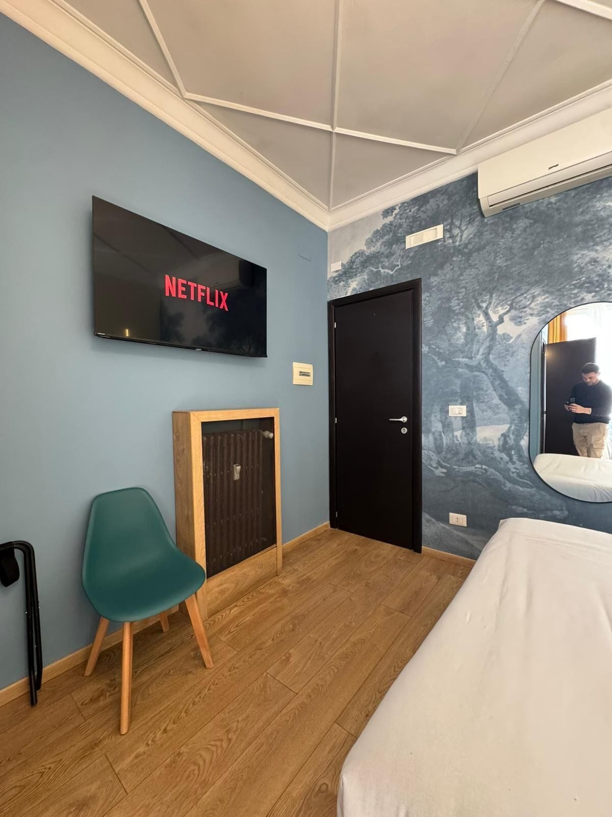 A room with a bed, a chair, a door, a wall-mounted TV displaying 'NETFLIX,' and a mural on the wall.