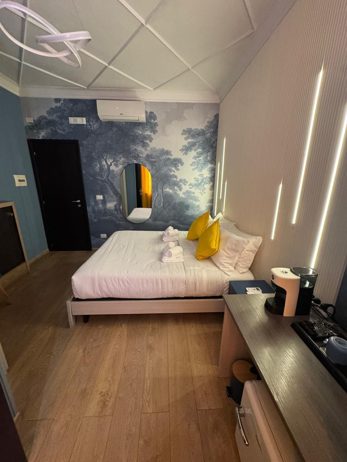 A modern hotel room with a bed, a desk, a mirror, and a wall with a tree mural.