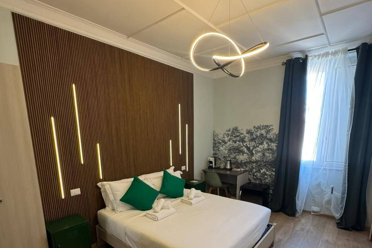 A modern hotel room with a wooden floor, a bed with white linens, green pillows, a desk with a chair, and a painting on the wall.