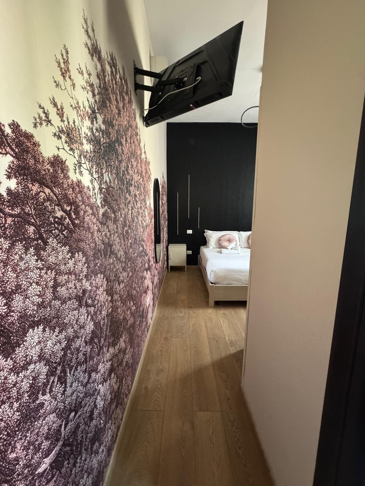 A narrow hallway with a bed, a wall-mounted TV, and a large tree mural.