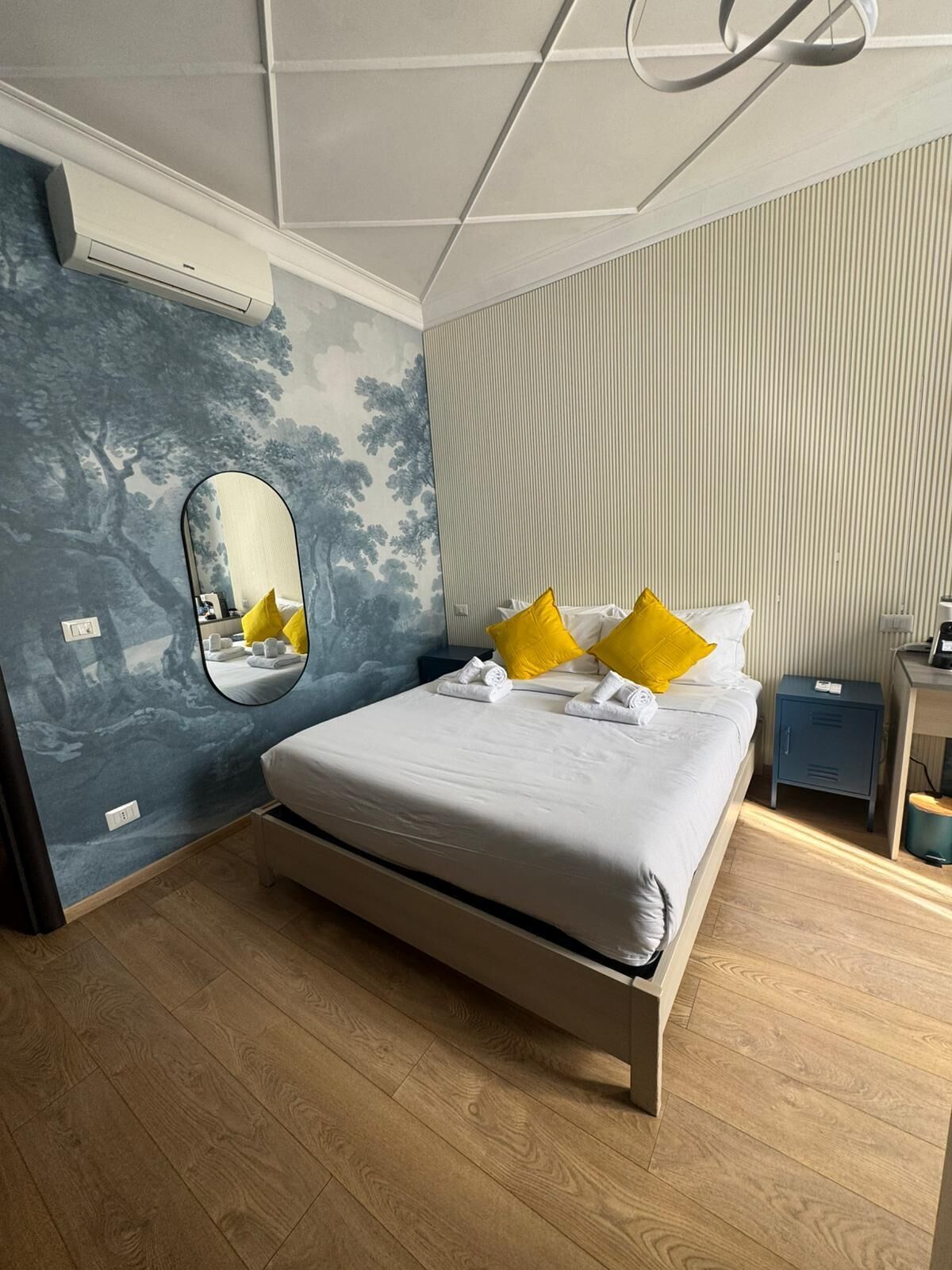 A bedroom with a bed, a mirror, a nightstand, and a wall with vertical lines.