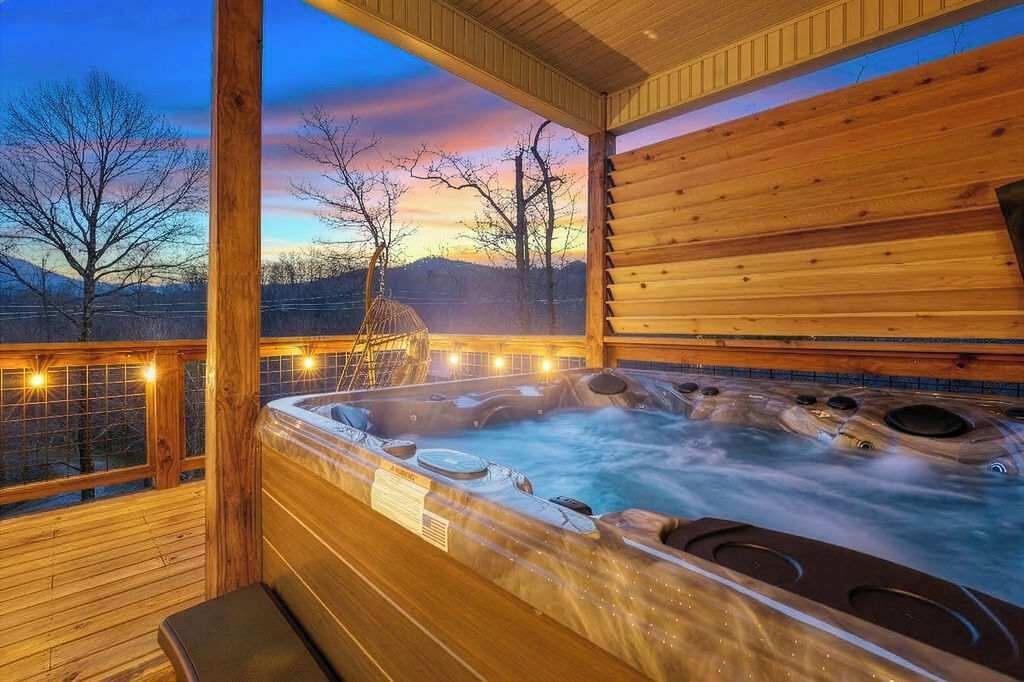 Un bain à remous doté de plusieurs jets, entouré d’une terrasse et de rampes en bois, offrant une vue sur les arbres et un coucher de soleil.