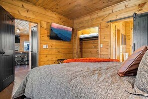 Ein Holzblockhaus-Interieur mit einem Bett, einem Gemälde an der Wand und einem Essbereich, der durch eine offene Tür sichtbar ist.