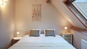 A bedroom with a sloped ceiling, a macrame wall hanging, two bedside lamps, a bed with white bedding, and a window with a view of the outside. - La Petite Histoire (Ath)