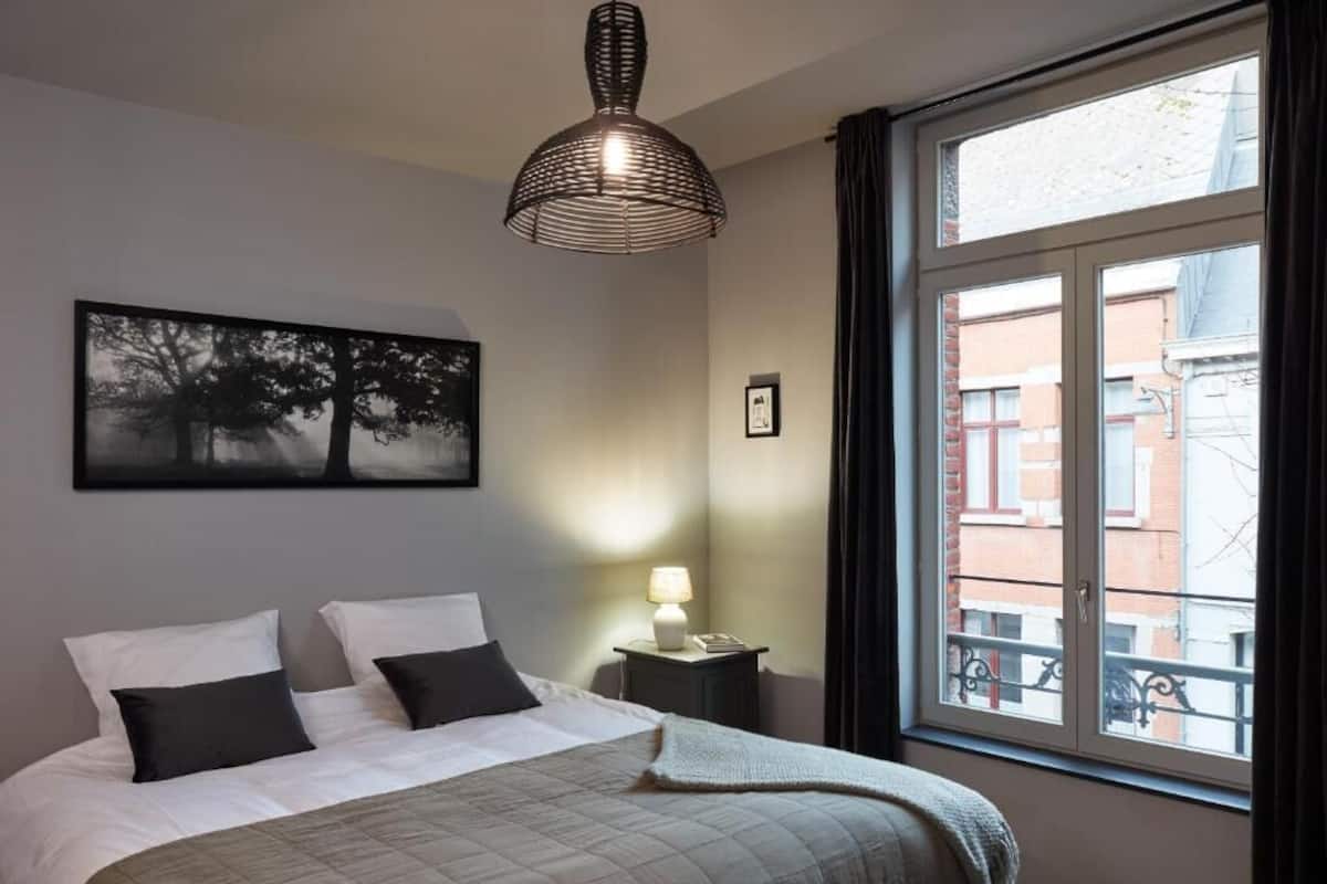 A bedroom with a bed, a bedside table with a lamp, a window with a view of buildings, and a wall-mounted artwork.