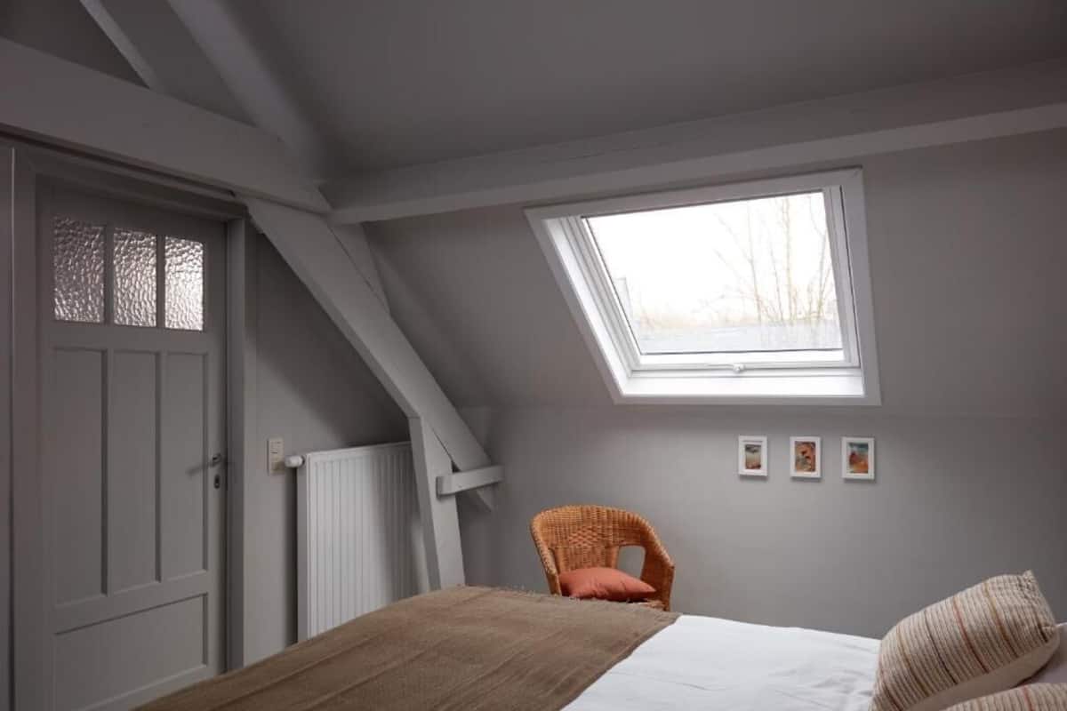 A bedroom with a sloped ceiling, a skylight, a bed with pillows, a chair, and a radiator.