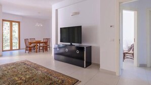 A modern living room with a flat-screen TV on a black cabinet, a dining area with wooden chairs, and a large patterned rug on the floor. - Cozy apartment in Lecce, parking (Lecce)