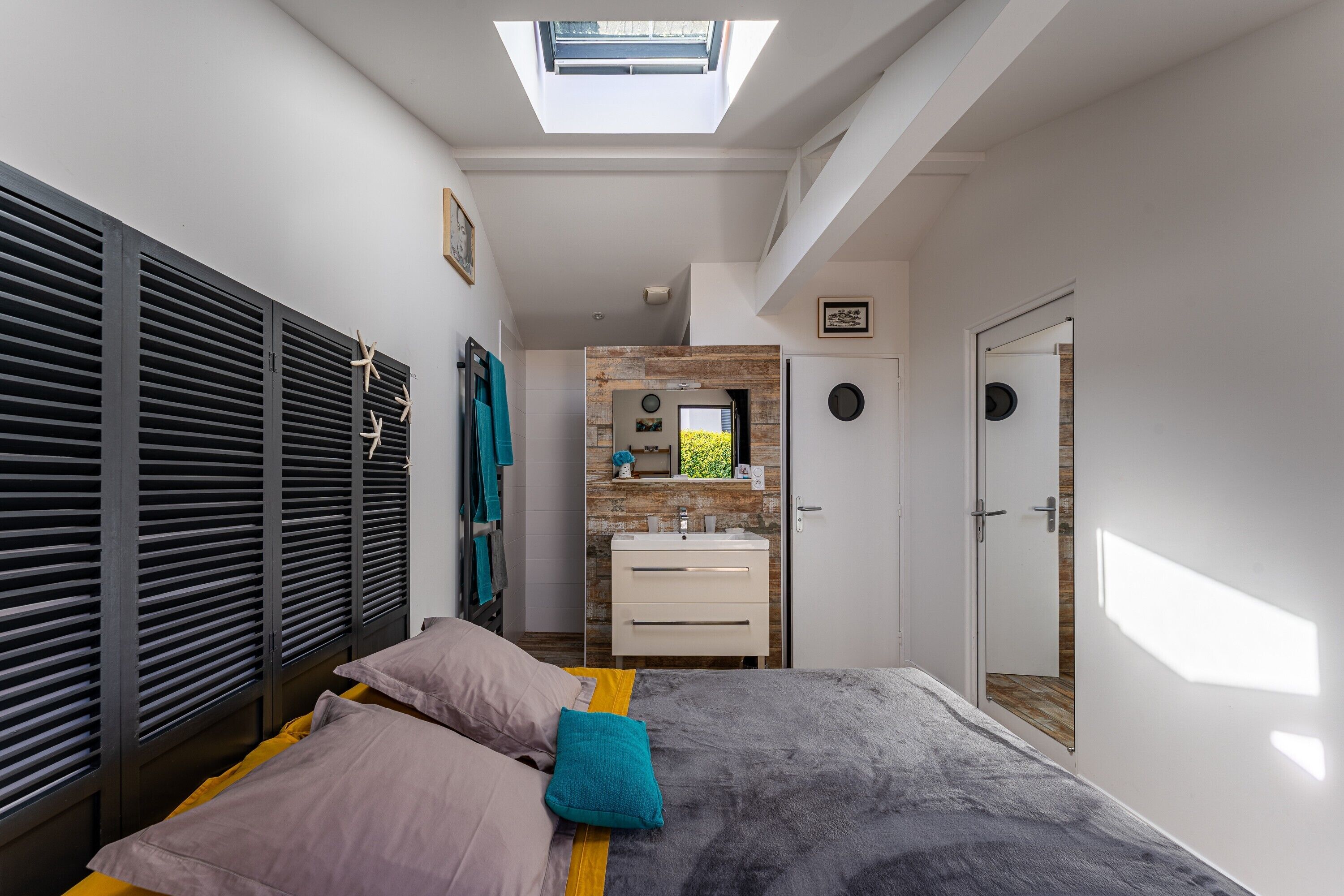 A bedroom with a skylight, a bed with pillows, a bedside table, a wardrobe with shutters, and a bathroom with a mirror and sink.