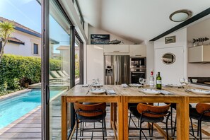 Comedor con vista a la piscina, mesa de madera, sillas y cocina equipada con electrodomésticos modernos.