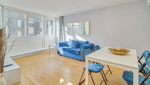 A modern living room with a blue sofa, a dining table, and a large window. - Central London Stay | Steps from Regent’s Park (London)
