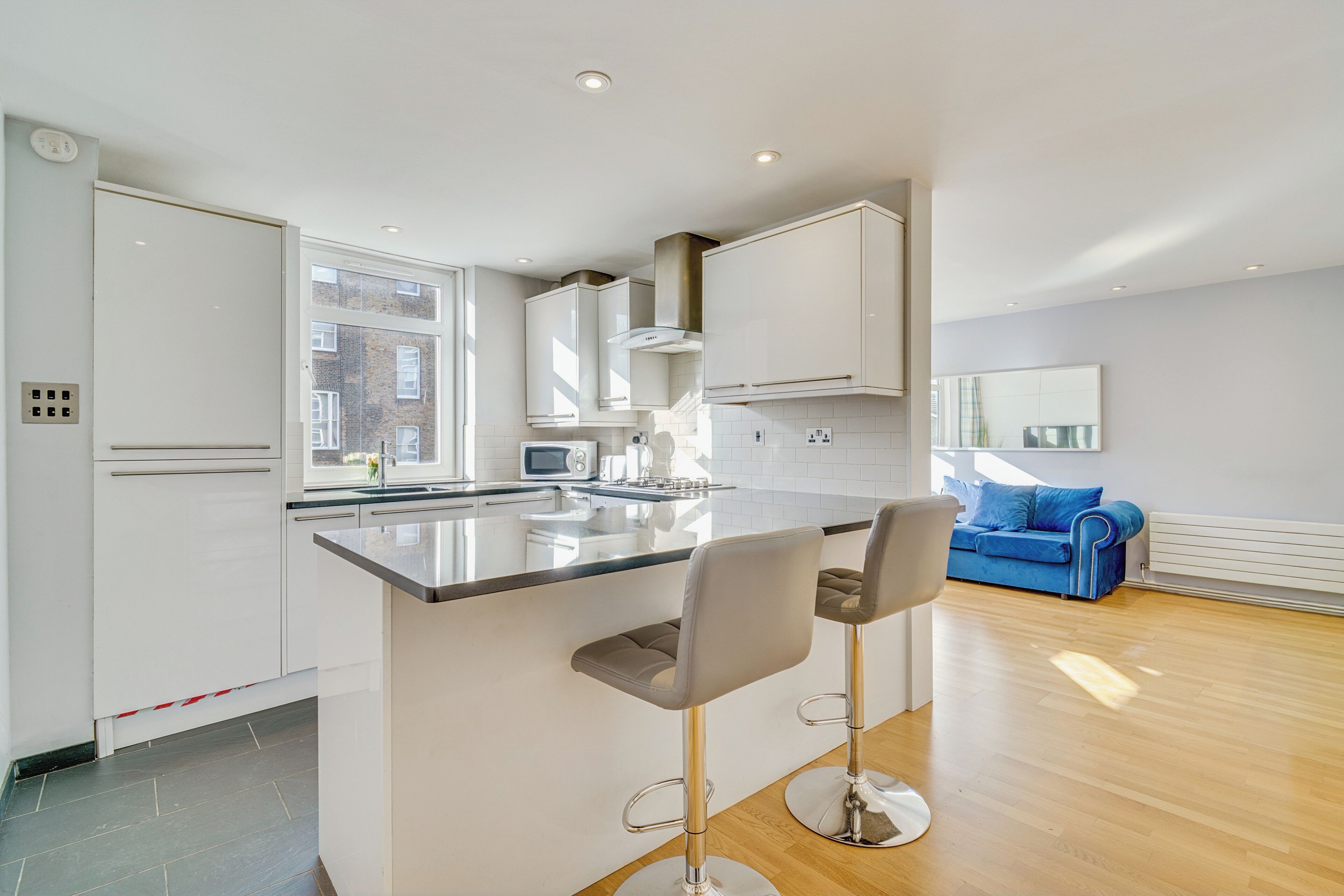 A modern kitchen with a central island, bar stools, a blue sofa, and a large window.