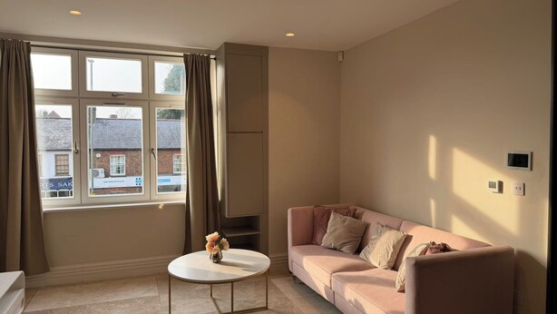A living room with a sofa, a small round table, and a window with curtains. - London 2 Bed Premium Flat (London)