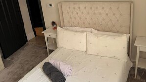 A neatly made bed with a tufted headboard, a bedside table with a mirror, and a door leading to another room. - London 2 Bed Premium Flat (London)