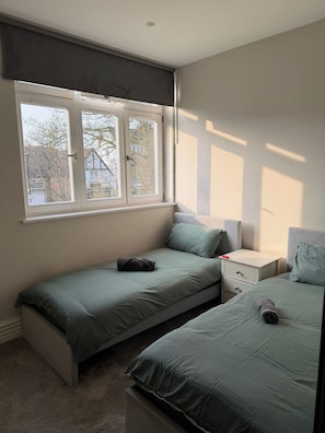 Ein Schlafzimmer mit zwei Einzelbetten, einem Nachttisch, einem Fenster mit Jalousien und Blick auf Bäume und Gebäude.