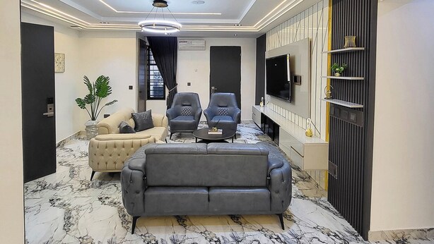 A modern living room with a sofa, two armchairs, a TV, and a decorative wall panel. - KELVIC SUITES (Owerri)
