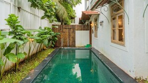 A swimming pool with a wooden fence, lush greenery, and a building with a tiled roof and a window. - ELIVAAS Amayah Vayu 6 BHK Villa (Assagao)
