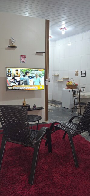 Une pièce avec un téléviseur diffusant un programme d’information, des fauteuils en osier noir, un tapis rouge, une petite table avec un panier et un mur recouvert de carreaux blancs.
