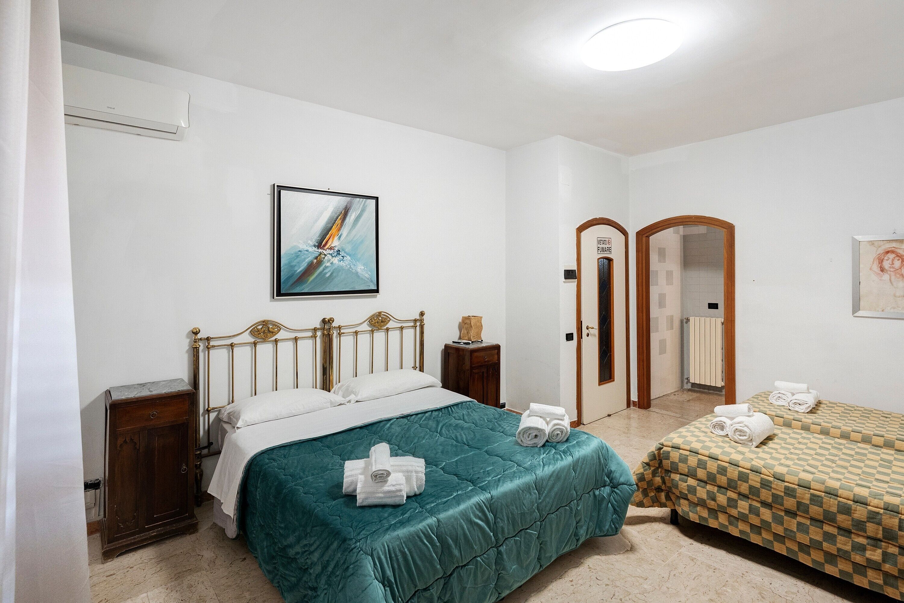Une chambre d’hôtel avec un lit, une petite table de chevet, une armoire, un tableau au mur et une porte donnant sur une autre pièce.