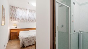 A bedroom with a bed, a nightstand, a window with floral curtains, and a shower area with a glass door. - H Pompeii Ruiins Apartment Hotel - Standard Double Room 4 (Pompei)