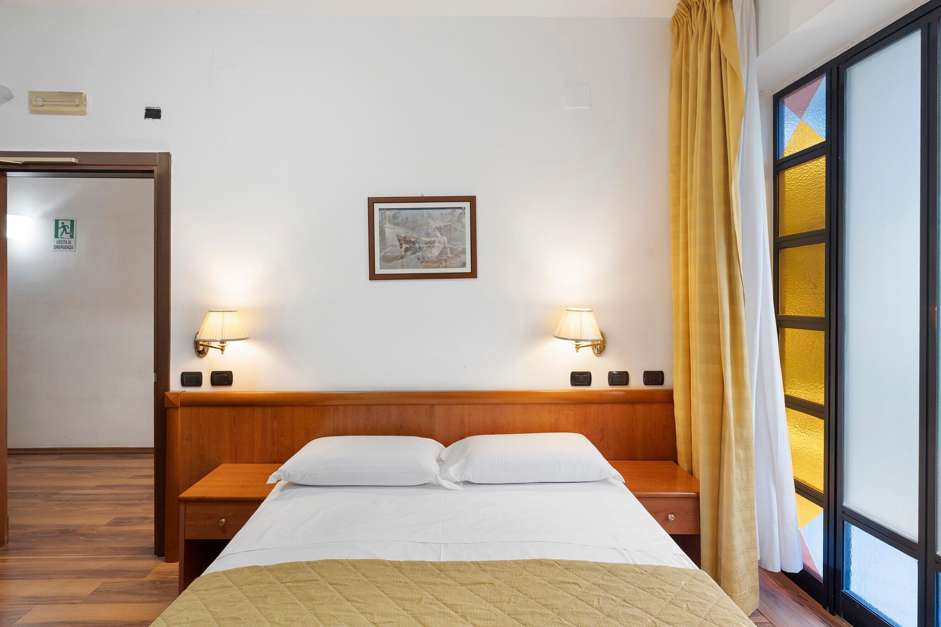 Een hotelkamer met een bed, bedlampjes, een foto in een lijst aan de muur, en een raam met gordijnen.