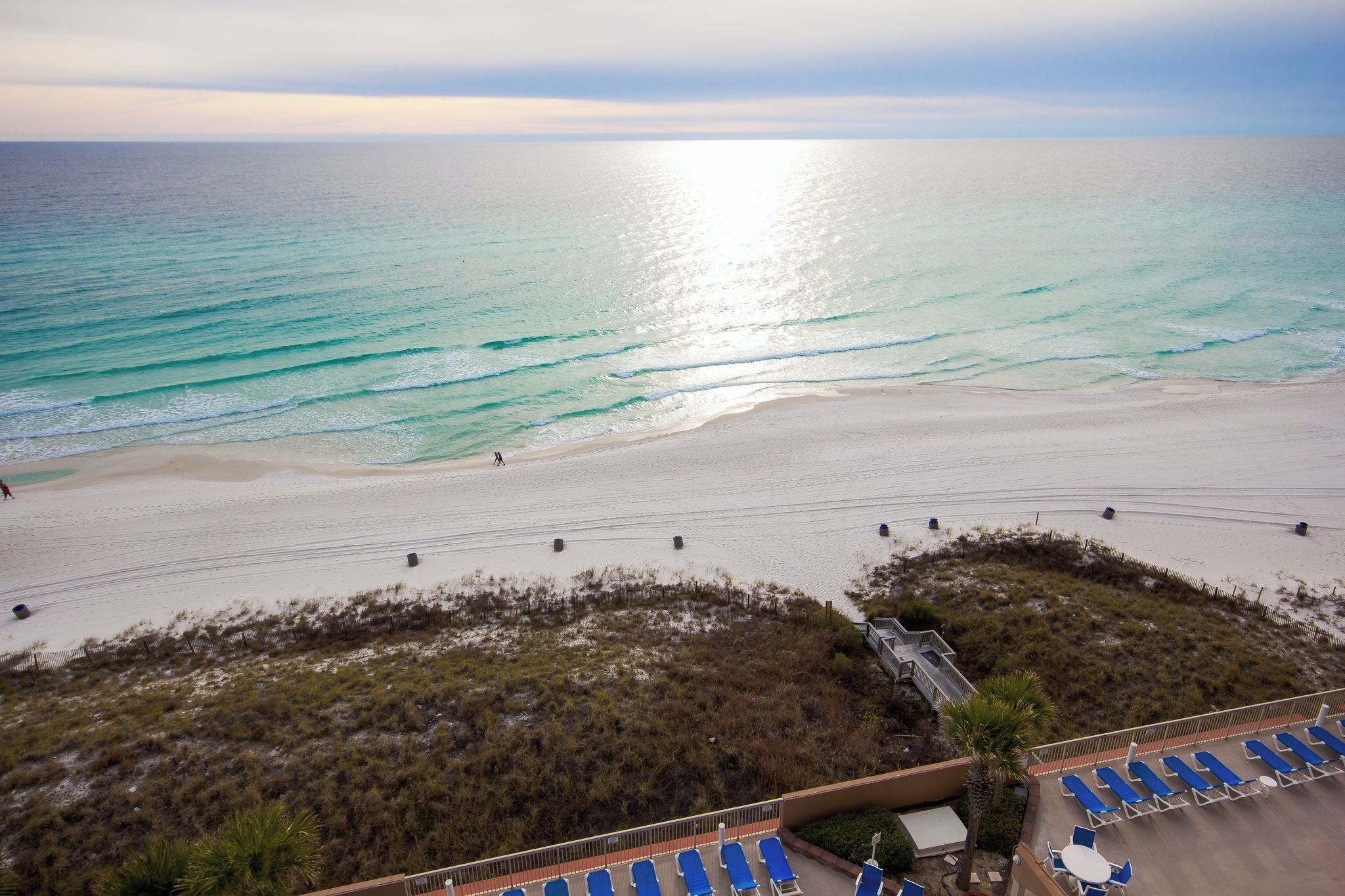 Blick auf einen Strand mit sandigem Ufer, ein paar Personen unterwegs und eine Reihe blauer Stühle.