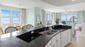 A modern kitchen with a large island, a dining area with chairs, and a view of the ocean. - Pelican Perch! 3BR 3BA Corner Beachfront Condo w/ Epic Views, Sleeps 10 (Panama City Beach)
