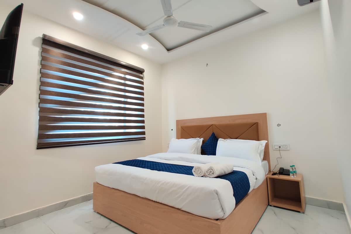A modern hotel room with a wooden bed, white bedding, a ceiling fan, a TV mounted on the wall, and a bedside table with a phone and water bottle.