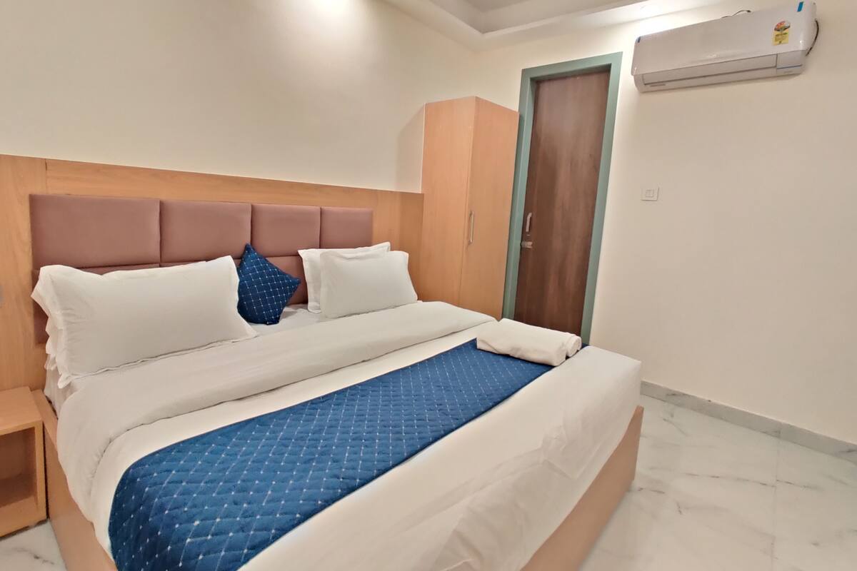 A neatly made bed with a blue quilt, a wooden headboard, and a white wall with an air conditioning unit.