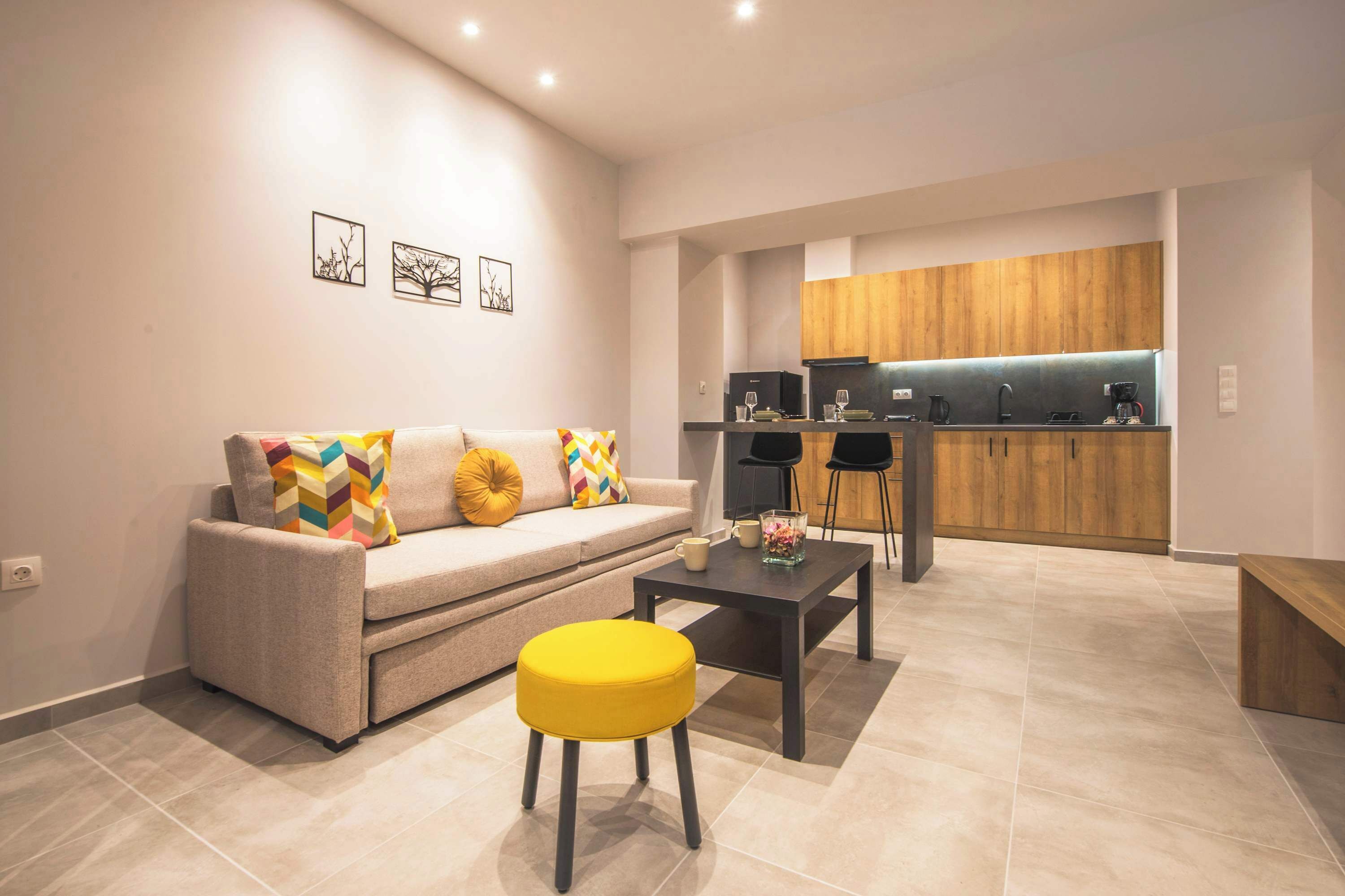 Una sala moderna con sofá, reposapiés y mesa de centro, una cocina con armarios de madera y una obra de arte en la pared.