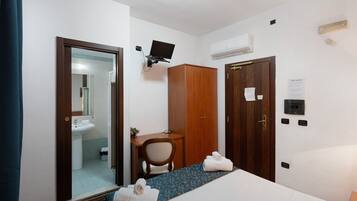 A hotel room with a bed, a desk, a chair, a wardrobe, a mirror, and a bathroom visible through an open door.