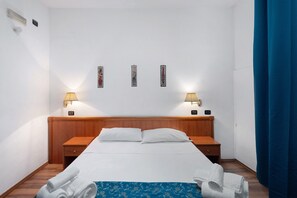 Een hotelkamer met een houten hoofdbord, twee nachtkastjes, een bed met witte lakens, twee wandlampen en een blauw gordijn.