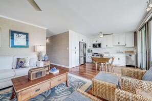 A modern living room with a sofa, coffee table, and wicker chairs. There is a kitchen area with white cabinets and stainless steel appliances. - Maui Parkshore #415 Ocean View in the Heart of Kihei, Walk to Kamaole Beach (Kihei)