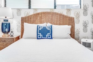 A neatly made bed with a woven headboard, a blue geometric pillow, and a white pillow. There is a wooden nightstand with a blue vase and a clock, and a white air conditioning unit beside the bed. - Maui Parkshore #415 Ocean View in the Heart of Kihei, Walk to Kamaole Beach (Kihei)
