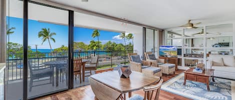 A spacious living area with a large sliding glass door leading to a balcony, furnished with a wooden table and chairs, a sofa, and a coffee table.
