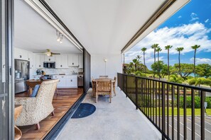A balcony with a dining table and chairs, a kitchen with white cabinets, and a view of palm trees and greenery. - Maui Parkshore #415 Ocean View in the Heart of Kihei, Walk to Kamaole Beach (Kihei)
