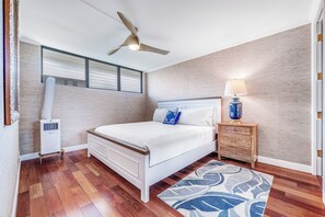 A bedroom with a bed, a nightstand, a lamp, a ceiling fan, and a window with blinds. - Maui Parkshore #415 Ocean View in the Heart of Kihei, Walk to Kamaole Beach (Kihei)