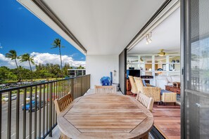 A balcony with a wooden table and chairs, a view of a parking lot, and a glimpse of the interior with wicker furniture and a ceiling fan. - Maui Parkshore #415 Ocean View in the Heart of Kihei, Walk to Kamaole Beach (Kihei)
