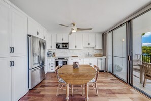 A modern kitchen with white cabinetry, stainless steel appliances, and a dining table with chairs. - Maui Parkshore #415 Ocean View in the Heart of Kihei, Walk to Kamaole Beach (Kihei)