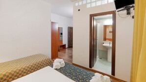 Habitación de hotel con cama, banca, espejo, televisión y baño a la vista a través de una puerta abierta.