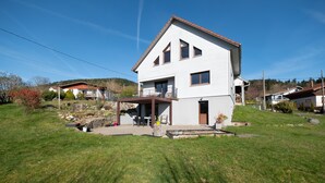 Une maison à deux étages avec un patio couvert, un petit jardin et un ciel d’un bleu éclatant.