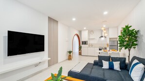 Una sala de estar moderna con un televisor de pantalla plana, un sofá oscuro con cojines, una mesa de centro de madera con una planta, y una cocina al fondo.