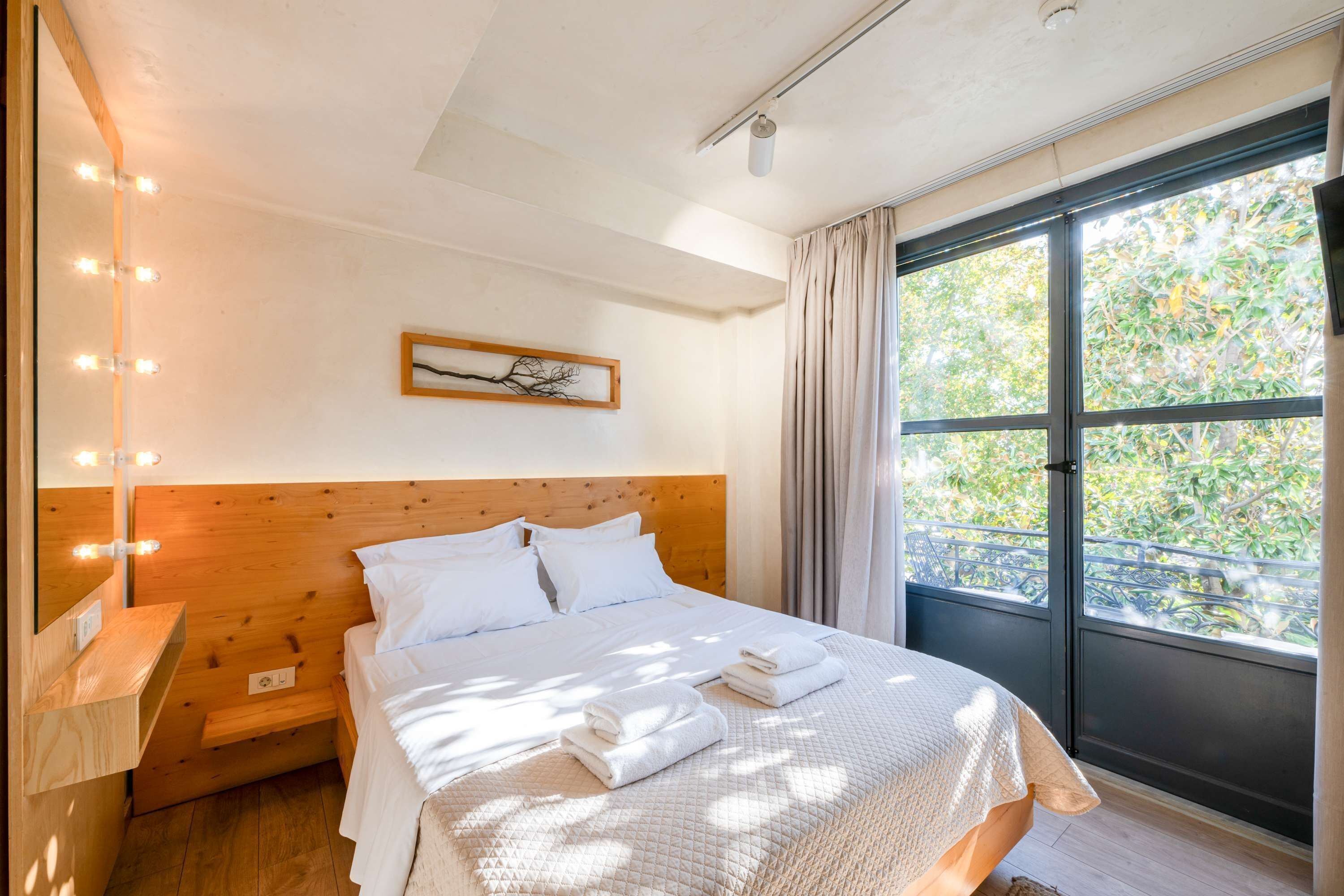 A bedroom with a wooden headboard, a large window with curtains, a bedside table with lights, and a framed picture on the wall.