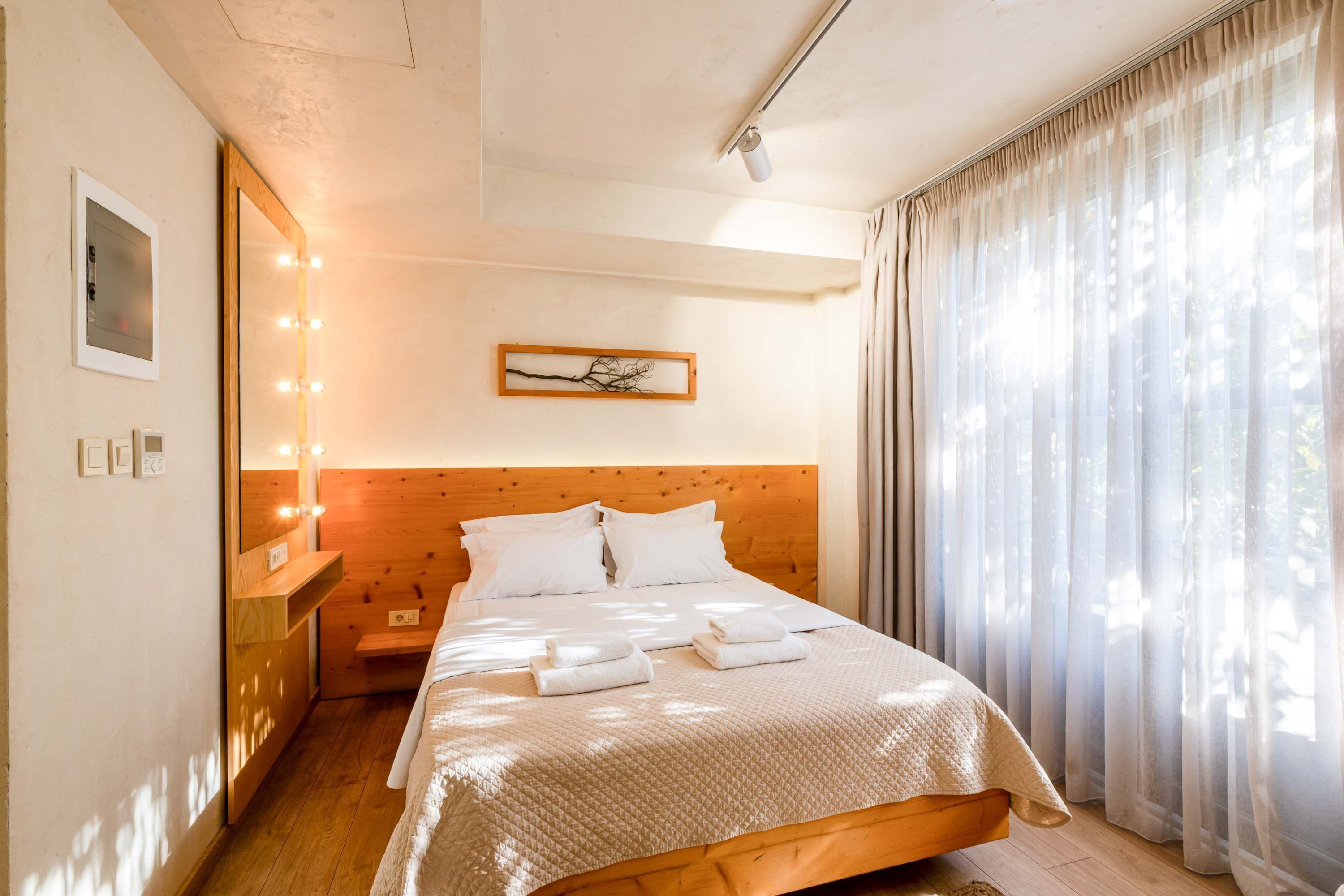 Une chambre d’hôtel avec une tête de lit en bois, un lit recouvert de draps blancs, une table de chevet avec une lampe et une fenêtre ornée de rideaux légers.